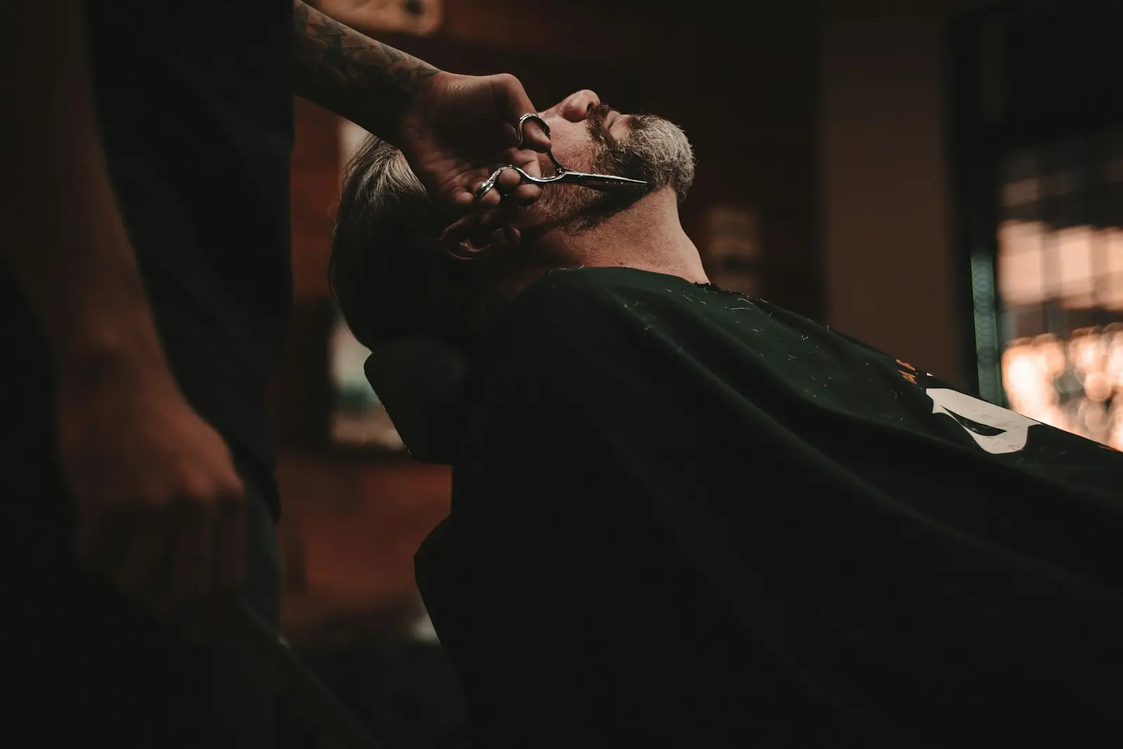 A barber trimming a customer's beard in a warmly lit shop
