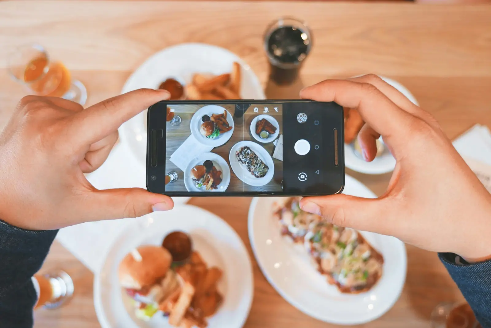 Two hands holding a phone over a table of plates, framing a photo of the food