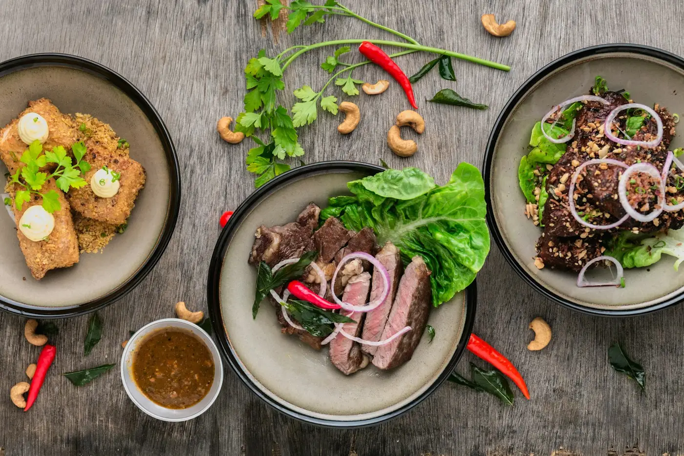 Overhead shot of three plated dishes with herbs, chillies and a dipping sauce on a wooden table