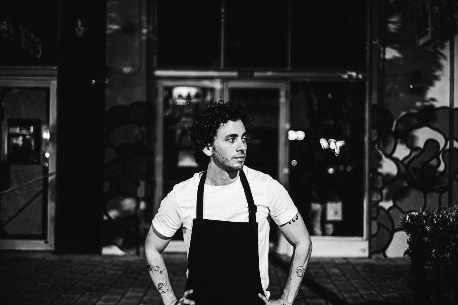 A café owner in a black apron standing outside his shop at night, hands on hips.