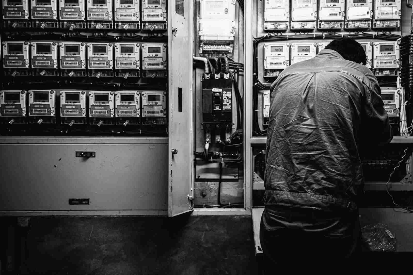 An electrician with his back to the camera, working at a wall of electricity meters.