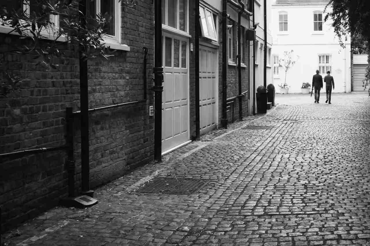 A narrow cobbled mews lane between brick buildings, two people walking away in the distance.