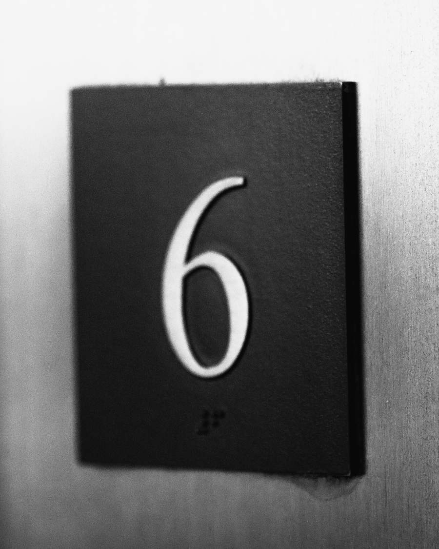 A black square plaque with the number six on a pale wall.