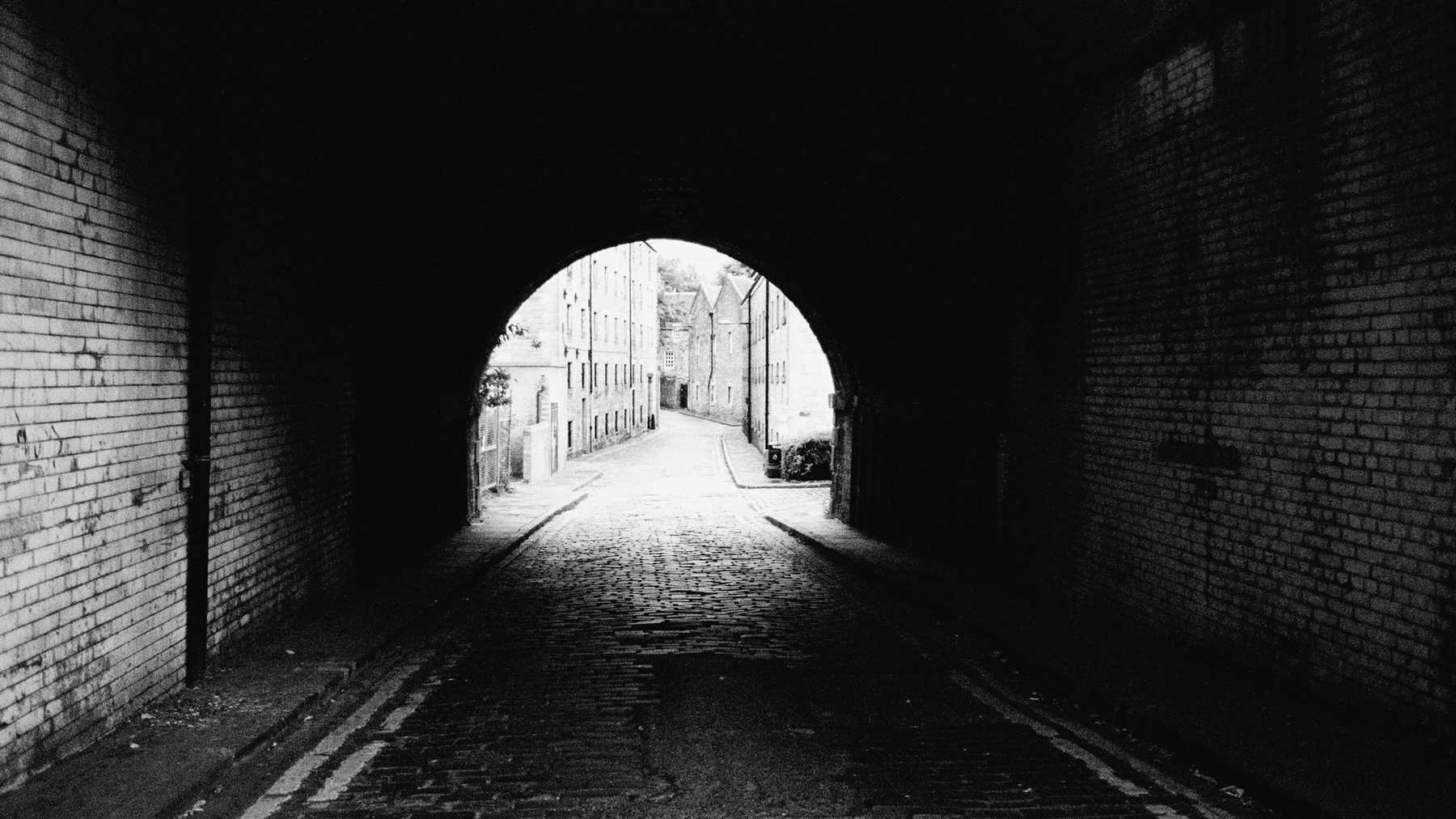 A long brick railway arch in black and white, dark inside, with a cobbled street in daylight at the far end.