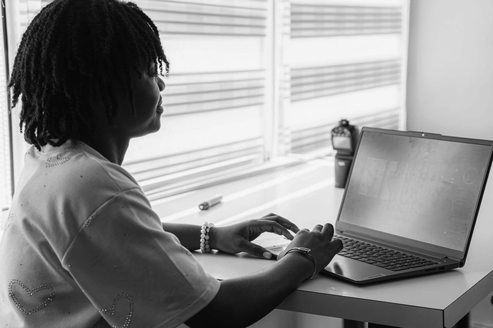 A self-employed designer typing on a laptop at a white desk by a window with blinds.