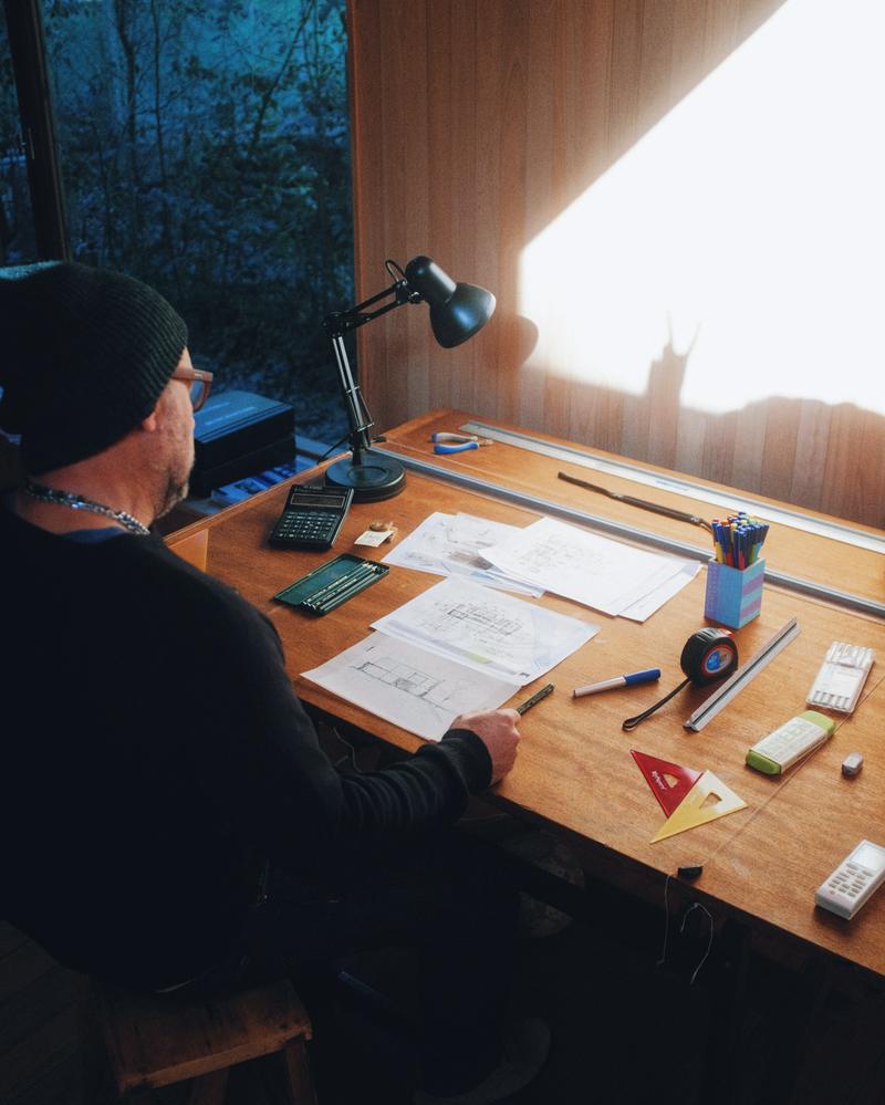 An architect working at a timber desk by a bright window, drawings spread out