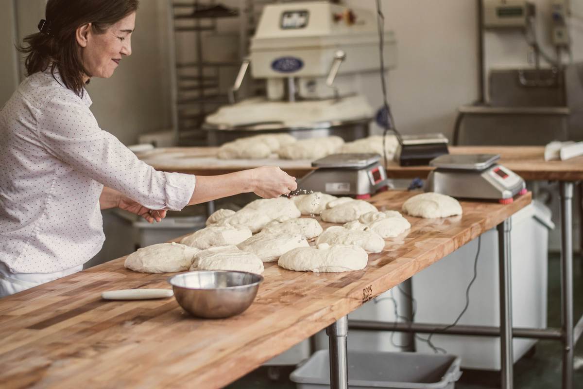 A baker dividing and shaping rounds of dough on a long wooden bench