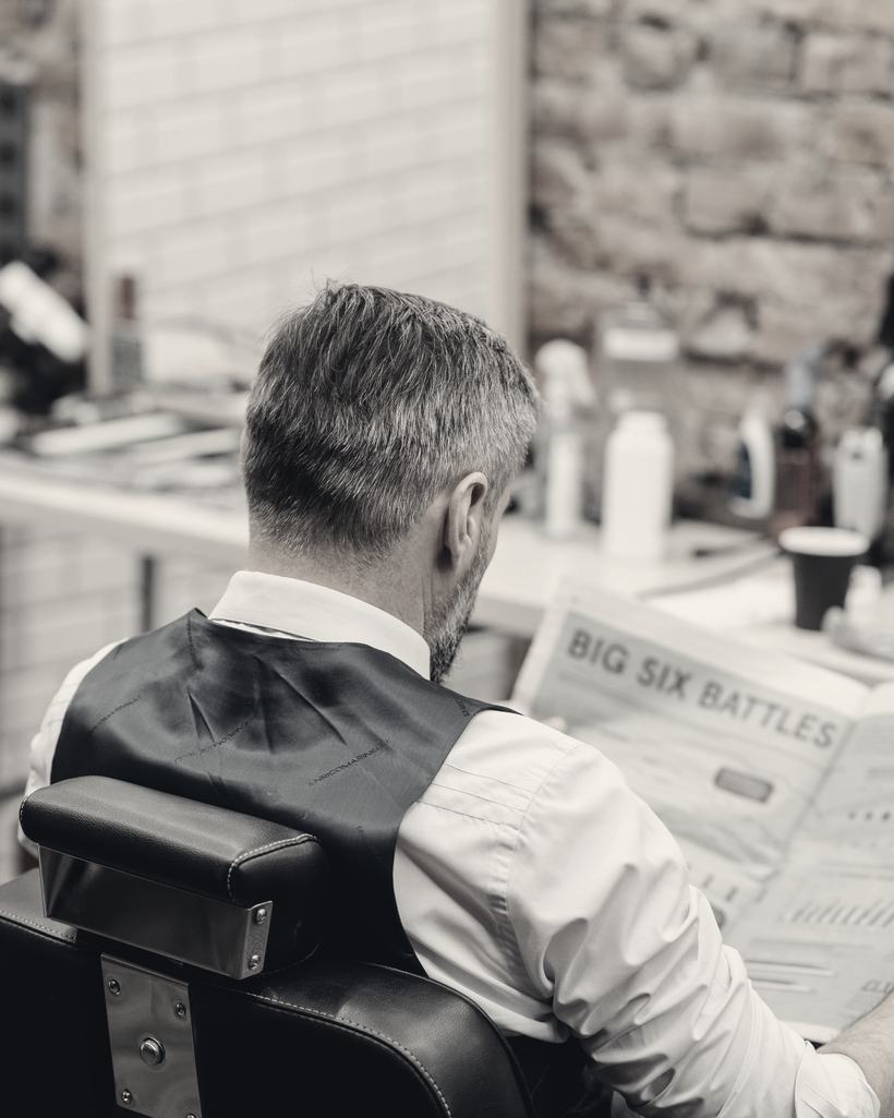 A client in a waistcoat reads the paper in a barber's chair before his cut