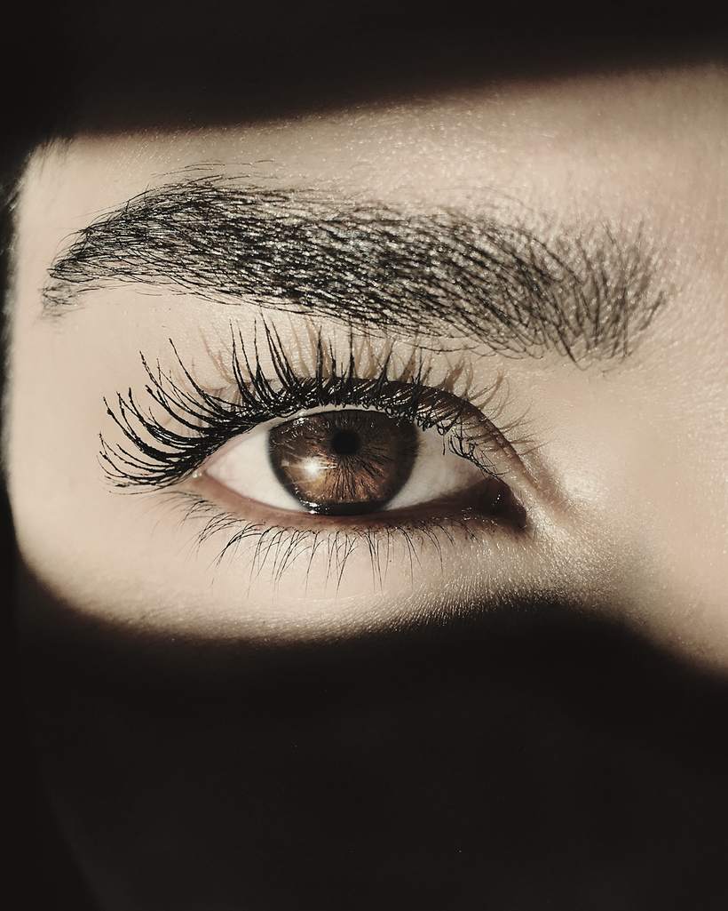 Close-up of a lashed eye and groomed brow lit by a narrow band of light