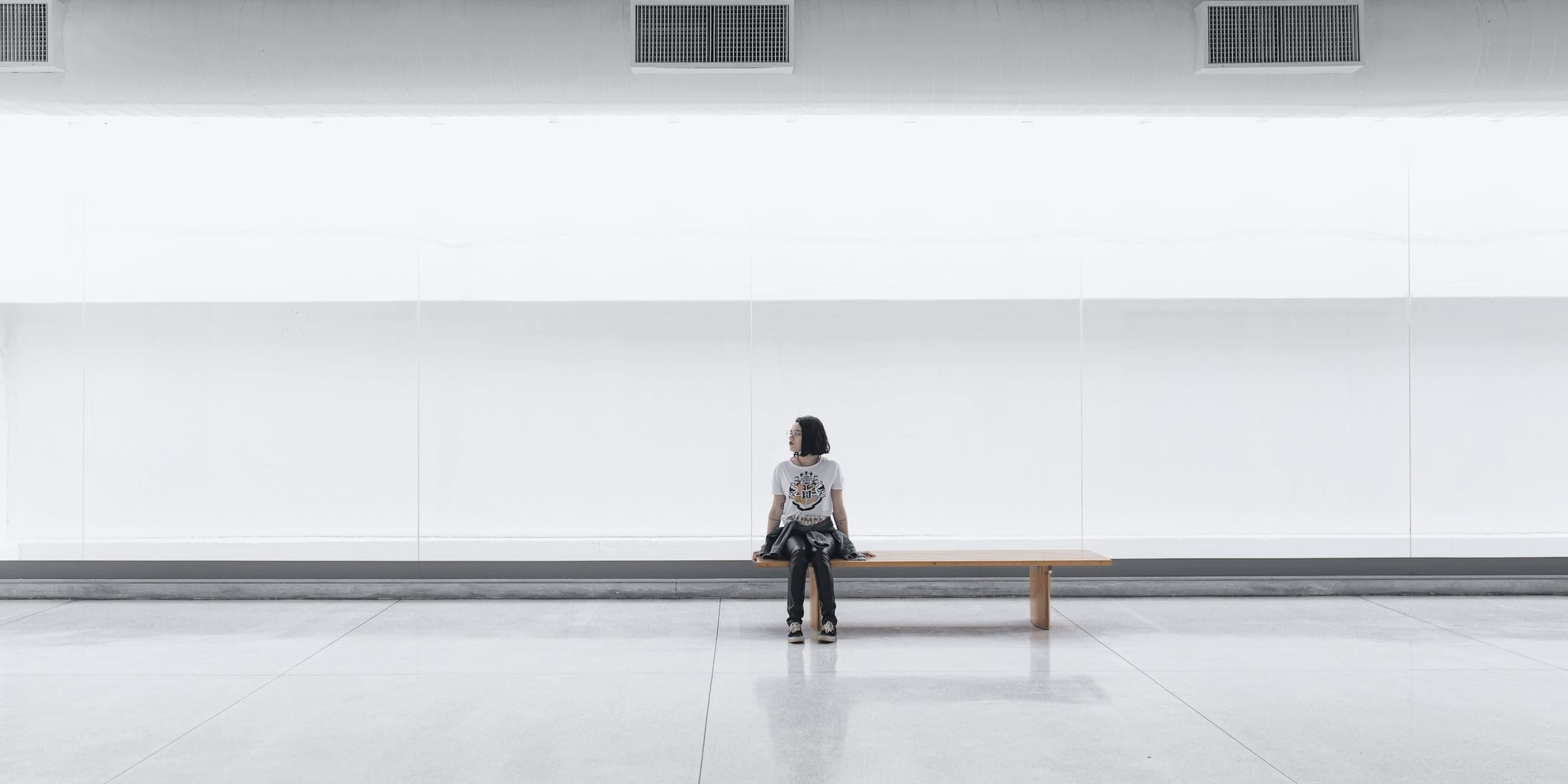A person sits alone on a long bench in a vast, bright white hall.