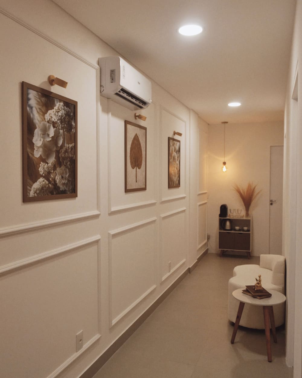 A calm clinic corridor with panelled walls, framed botanical prints and an armchair beneath a warm lamp.