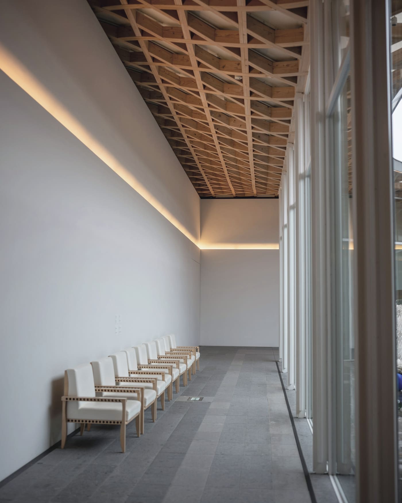 A long, quiet waiting gallery with a row of pale wooden chairs, daylight falling along a white wall beneath a timber lattice ceiling.