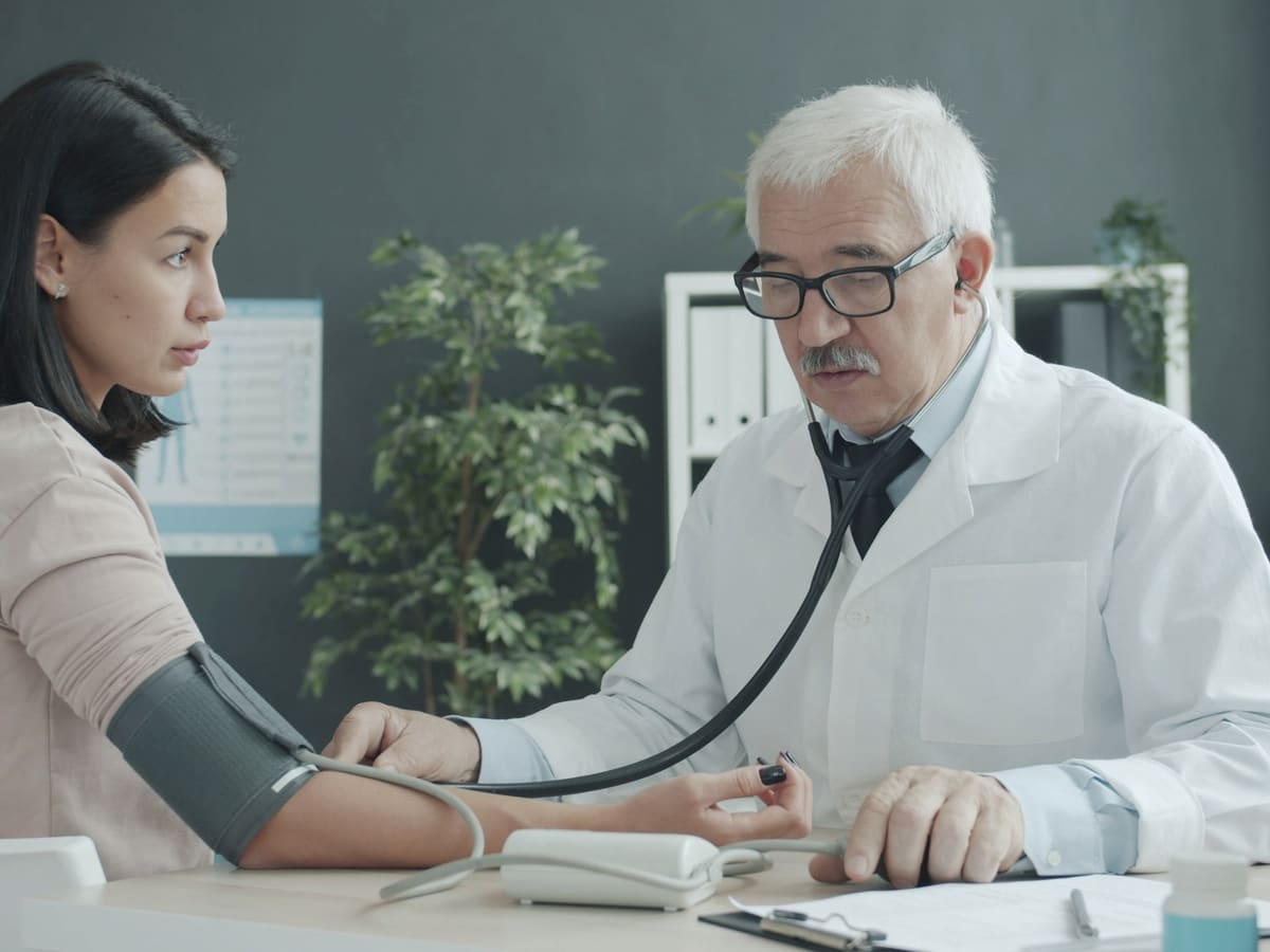A doctor takes a woman’s blood pressure with a cuff and stethoscope at a consulting-room desk.