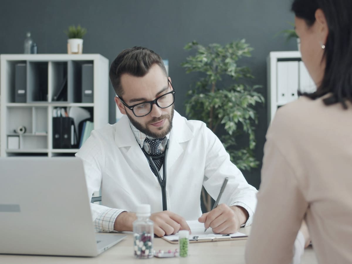 A doctor in a white coat writes notes at his desk while a patient sits opposite him.