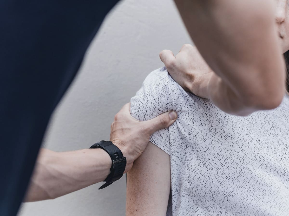A physiotherapist’s hands assess the shoulder of a patient in a grey T-shirt.