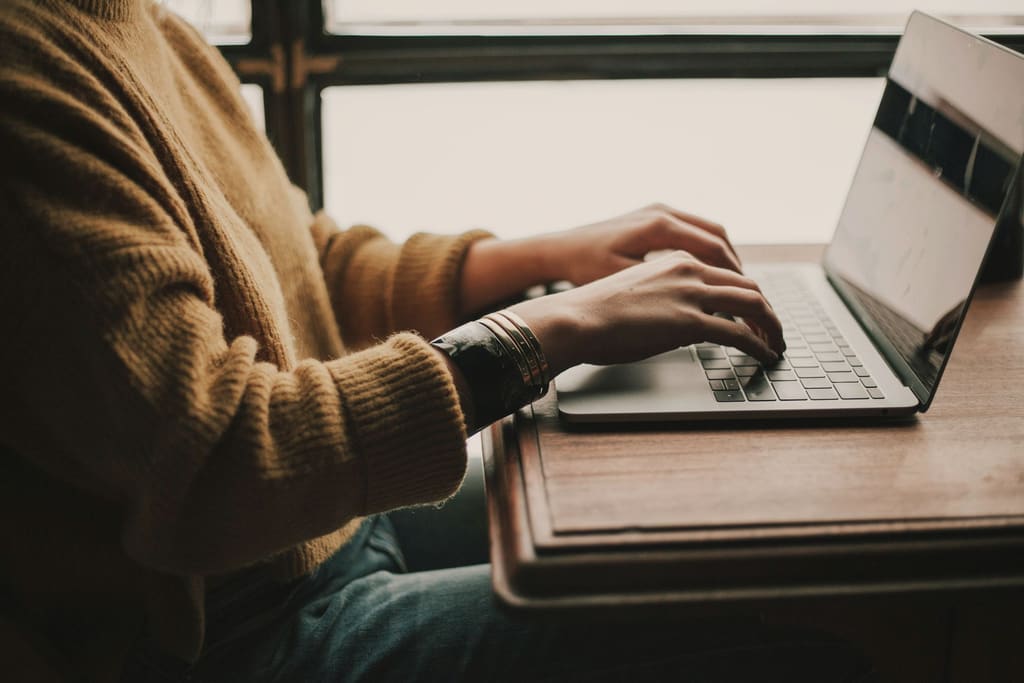 Someone in a mustard jumper typing on a laptop on a wooden table