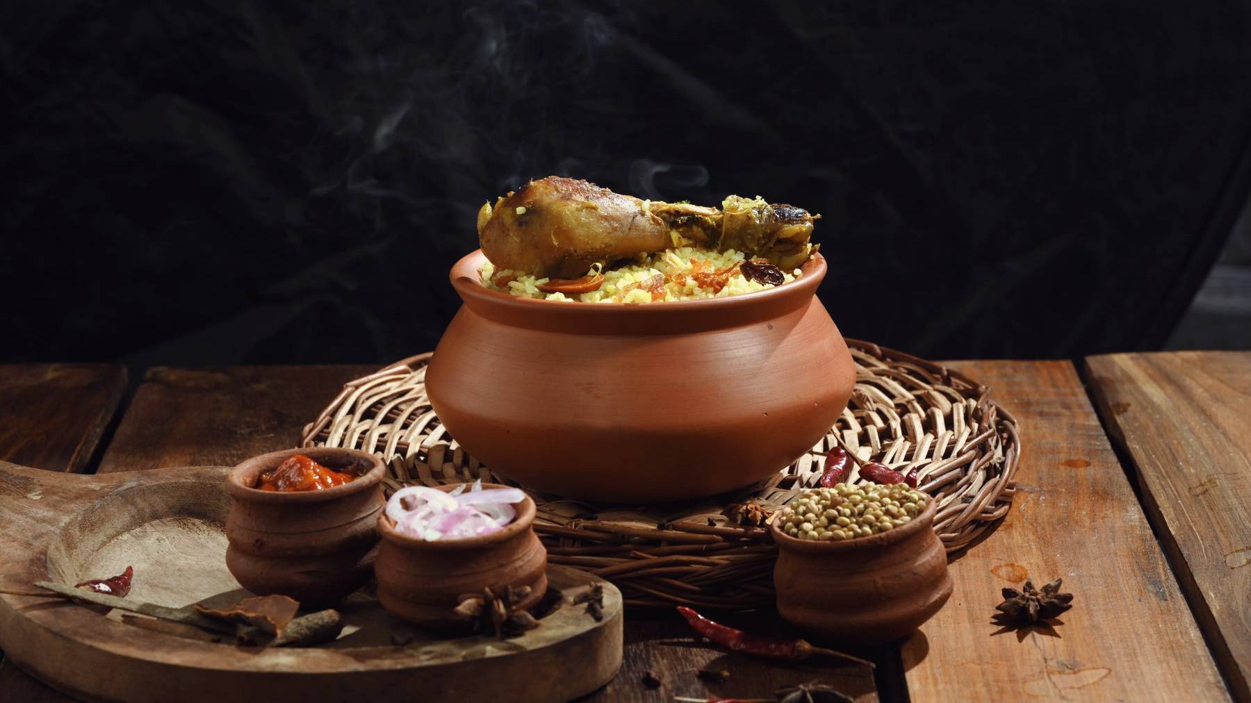 Chicken biryani sealed in a clay handi on a wicker mat, small bowls of spice beside it