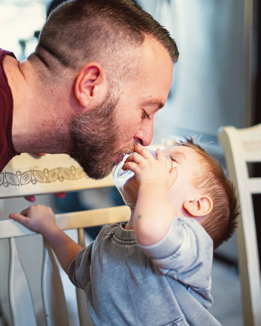 A dad kisses his toddler son on the head while the boy brushes his teeth