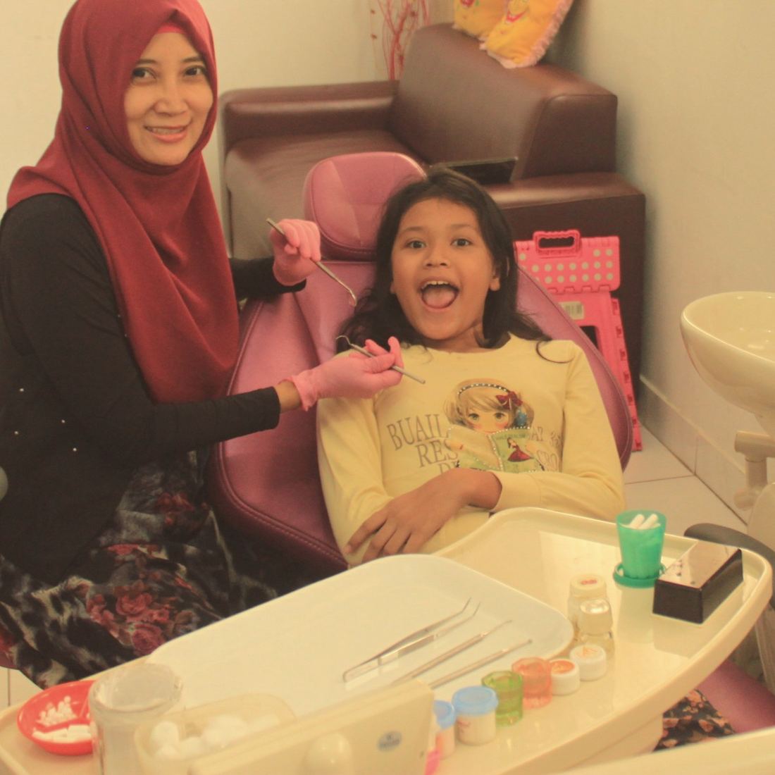 A dentist in a headscarf checks a smiling girl's teeth while she sits in a child-friendly dental chair