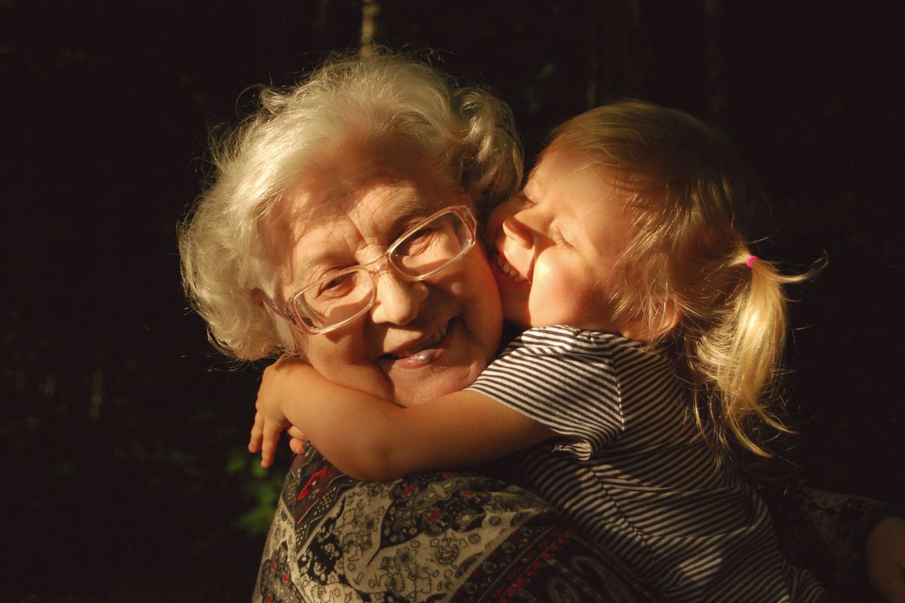 A grandmother laughing as her granddaughter hugs her tightly, in warm evening light