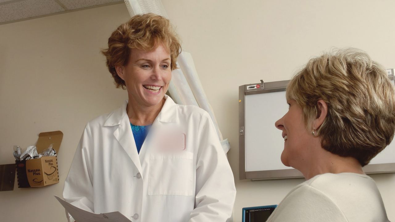 A smiling dentist holding a clipboard talks with a patient in a bright consultation room