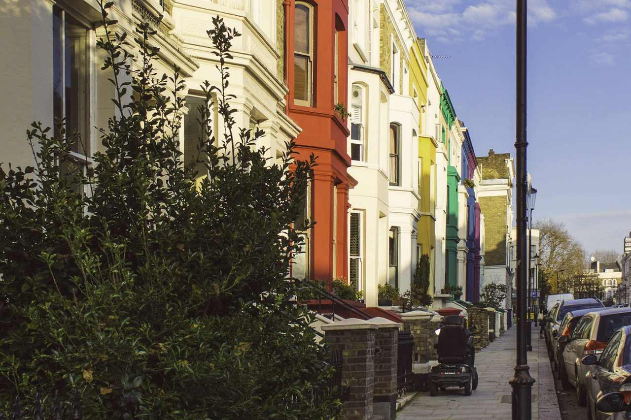 A row of brightly painted terraced houses on a sunny residential street, with a lamp post and a hedge