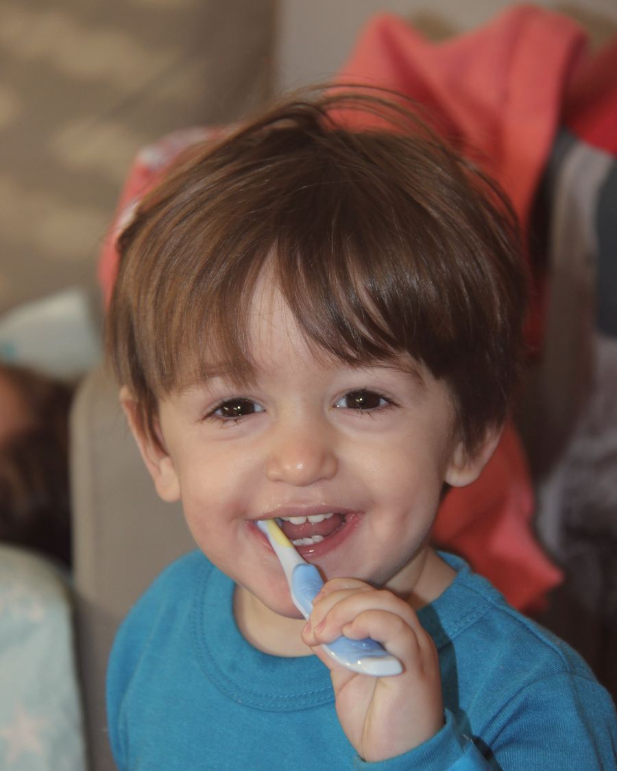 A toddler in a blue top grinning with a toothbrush in his mouth