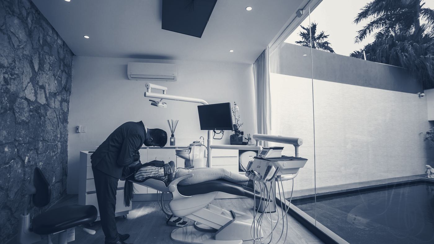 A dentist in a dark suit leans over a reclined patient in a stone-walled surgery that opens onto a courtyard pool