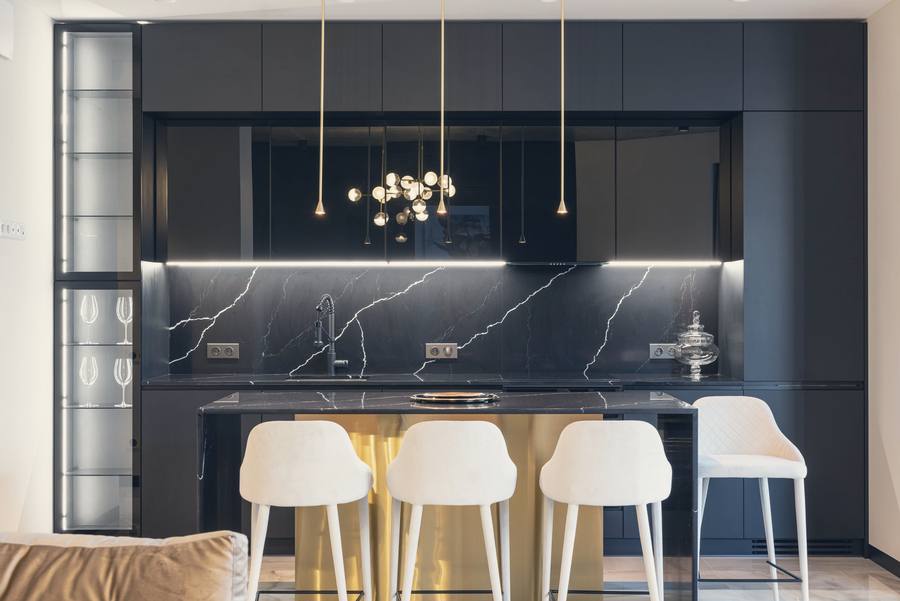 A contemporary kitchen with black cabinetry, a marble splashback, brass pendant lights and white bar stools
