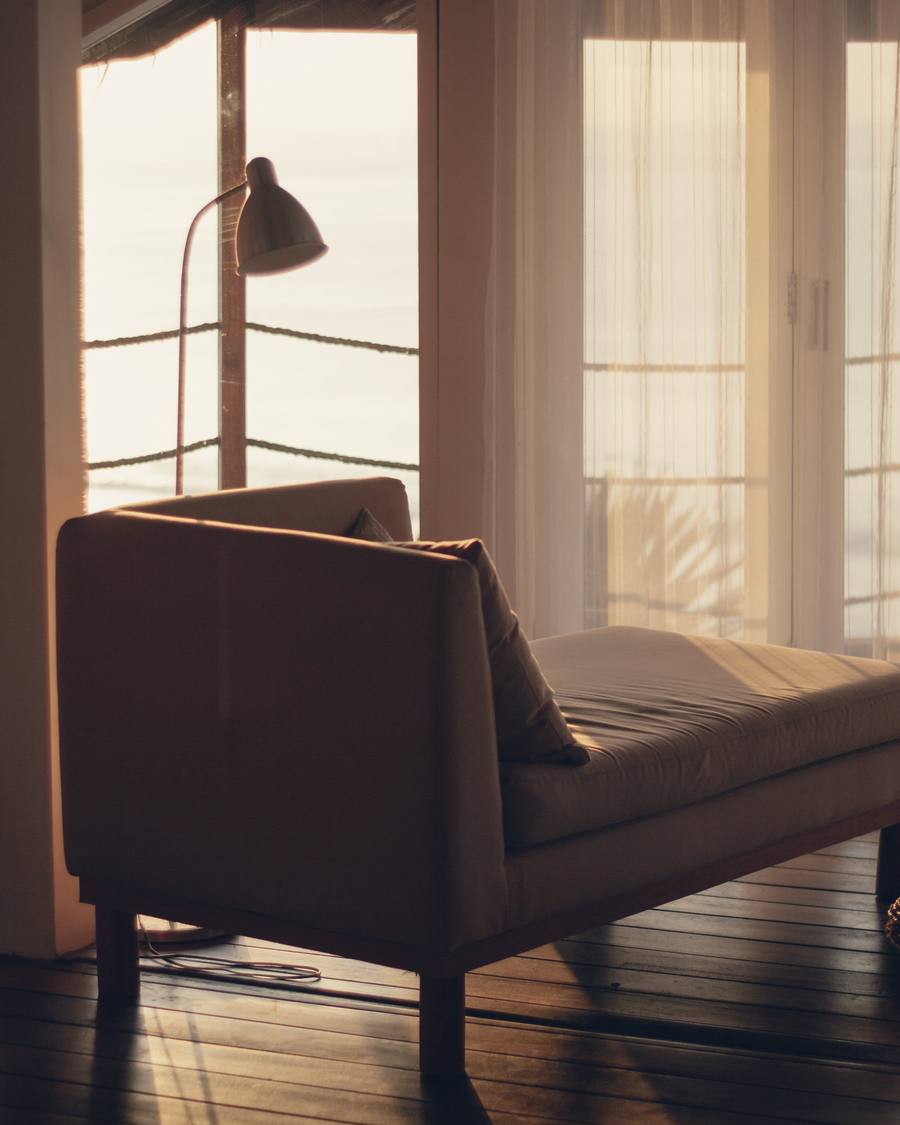 A quiet sitting room with a linen armchair and floor lamp in soft afternoon light behind sheer curtains