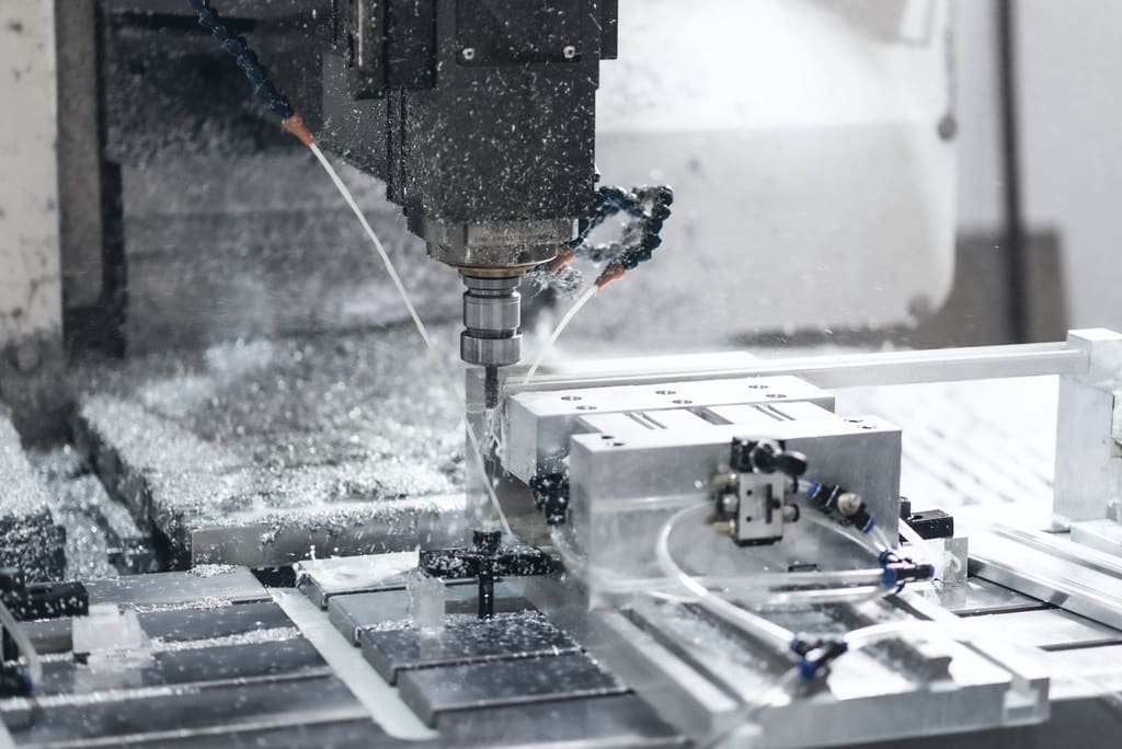 A vertical machining centre milling an aluminium block, coolant lines trained on the cutter and swarf across the table