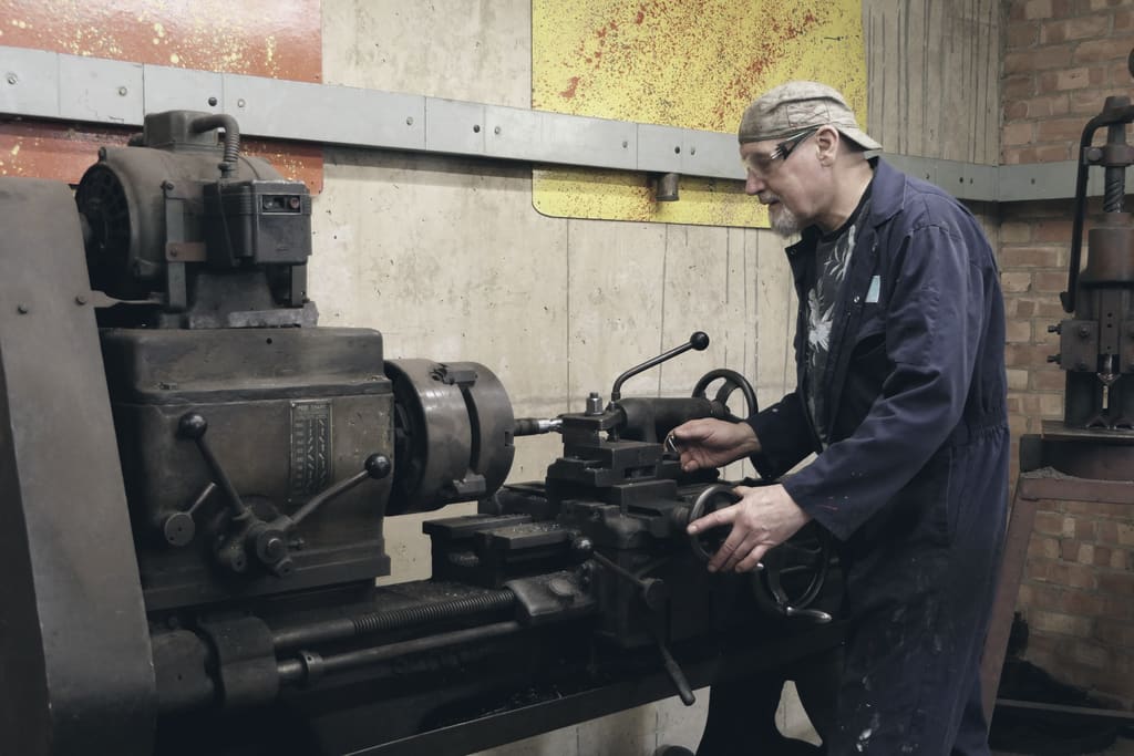 A machinist in overalls working at a manual lathe in a brick-walled workshop