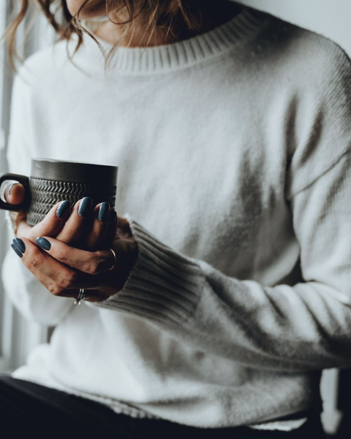 A woman in a cream knitted jumper holding a dark ribbed stoneware mug in both hands