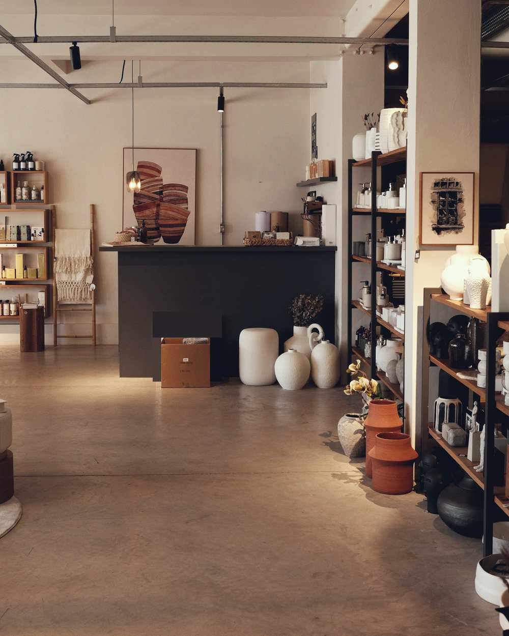 Inside a calm homeware shop: a long counter, white vases on a concrete floor, shelves of ceramics and a throw over a ladder