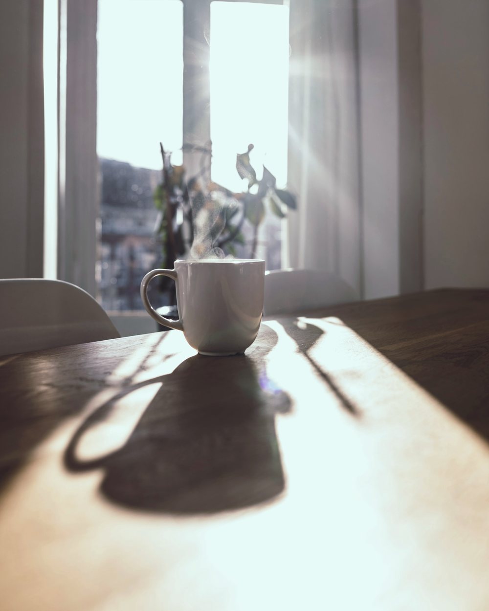A pale stoneware mug steaming on a wooden table in bright low morning sun