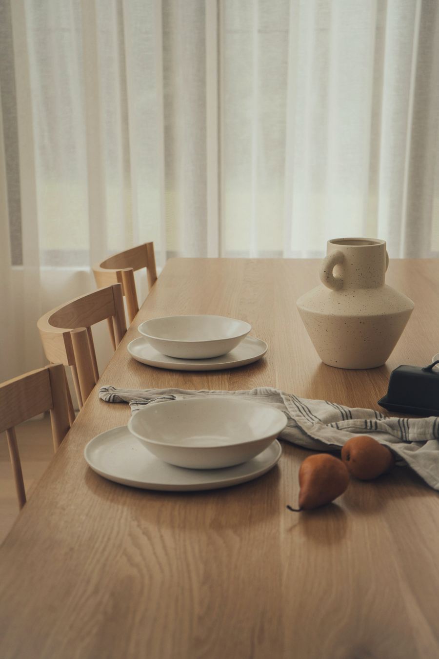 A wooden dining table by a curtained window, set with white bowls on plates, a speckled stoneware jug and a striped linen cloth