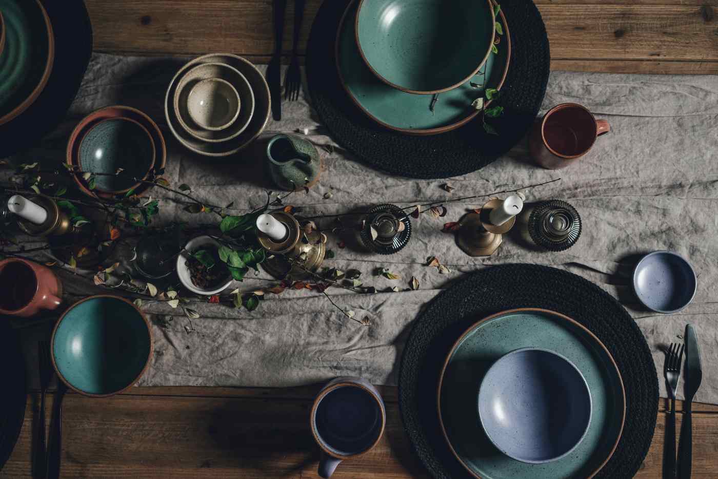 A long table seen from above: green and blue stoneware plates and bowls, candles in brass holders and a crumpled linen runner