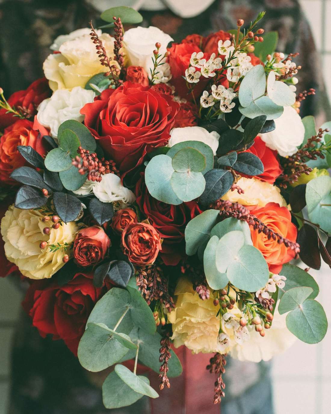 A florist holding a huge bouquet of red, peach and cream roses with eucalyptus against white tiles
