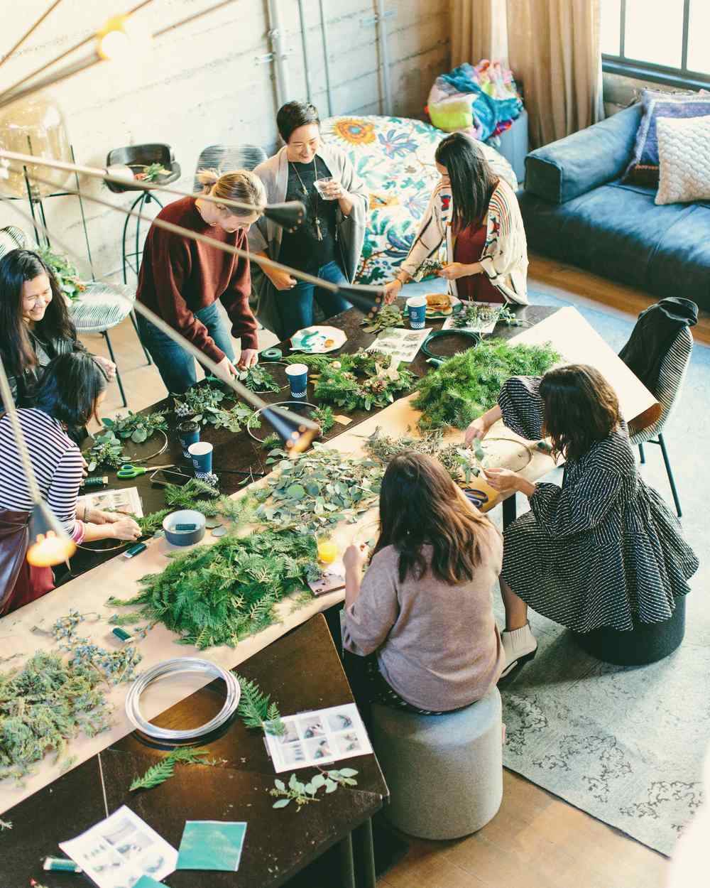 A group of people making wreaths together around a long table covered in greenery