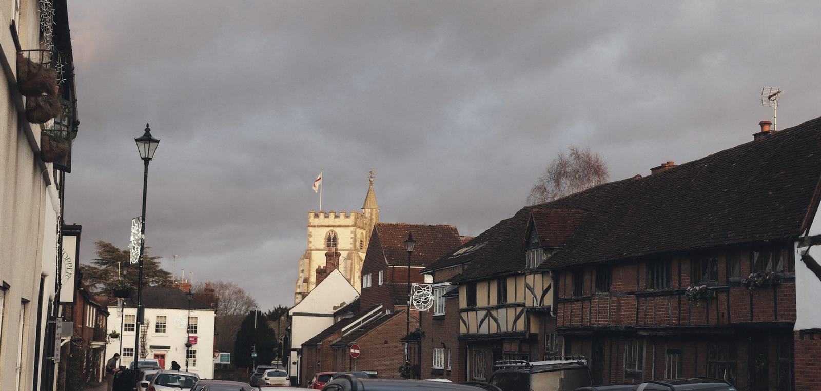 Timber-framed and brick houses along a market-town street at dusk, with a church tower under a heavy evening sky