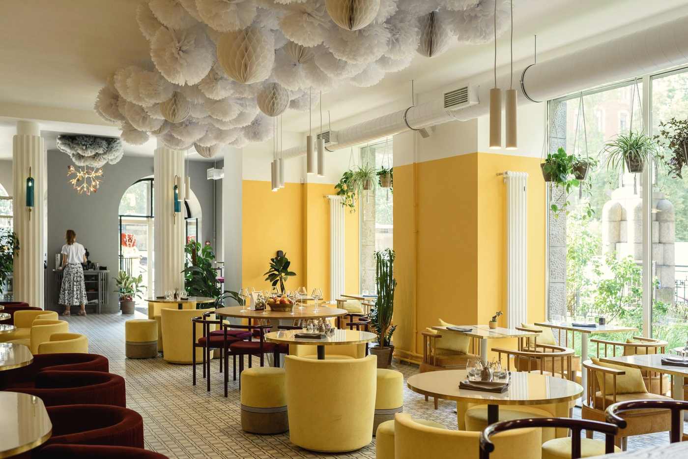 A bright dining room with yellow walls, round tables, velvet armchairs and hanging plants by tall windows