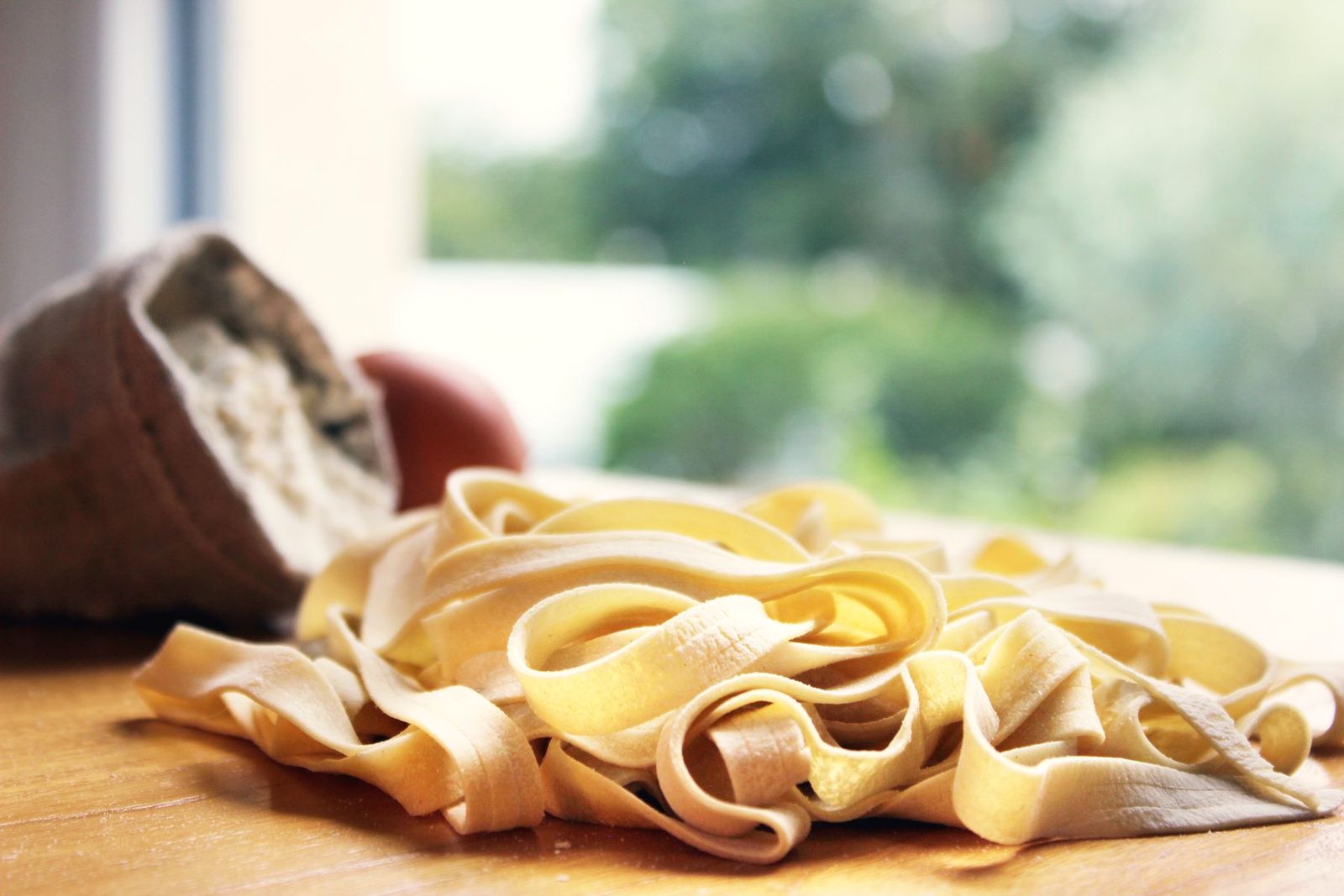 Ribbons of fresh egg tagliatelle on a wooden board in morning window light, a sack of flour behind