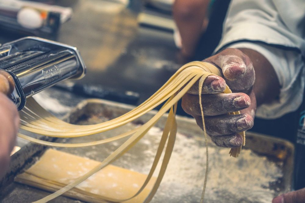 Strands of fresh pasta being fed through a hand-cranked pasta machine on a floury bench