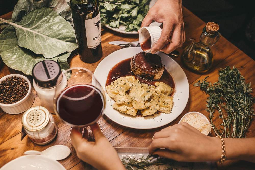 Hands around a wooden table pouring olive oil over a plate of gnocchi beside glasses of red wine